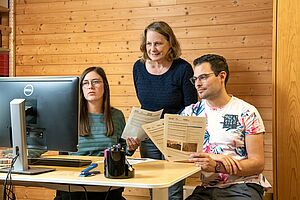 Trois collaborateurs devant leur ordinateur, un article à la main.