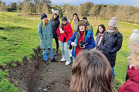 A woman with a camera speaks to a group next to an open area of ground. The participants are standing in warm clothing on a green meadow.