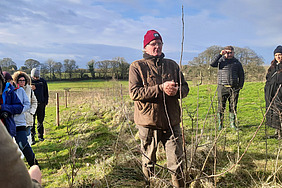 An elderly man in a red cap explains something next to a young tree in the field. Several warmly dressed people listen to him. 