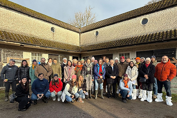 Group photo of 35 people in front of a building with a moss roof. They are dressed warmly, some are wearing shoe covers. The atmosphere is friendly and communal.