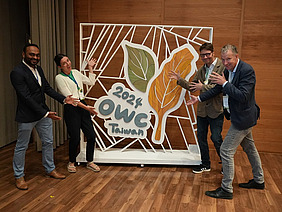 Four people pointing to a large OWC logo.