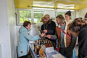 Auf einem Tisch stehen verschiedene Essigflaschen, drum herum stehen rund 6 Menschen.