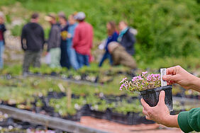 Eine Person hält einen kleinen Pflanztopf in die Kamera, im Hintergrund steht eine Gruppe Menschen.