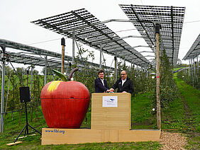 Eine hölzerne Bühne steht vor einer Niederstamm-Apfelanlage, auf welcher auf Metallstützen Photovoltaikmodule installiert sind. Auf der Bühne stehen ein grosser roter Styropor-Apfel und zwei Männer in bestem Alter im Anzug, welche einen grossen roten Knopf drücken, der symbolisch die Anlage eröffnet.